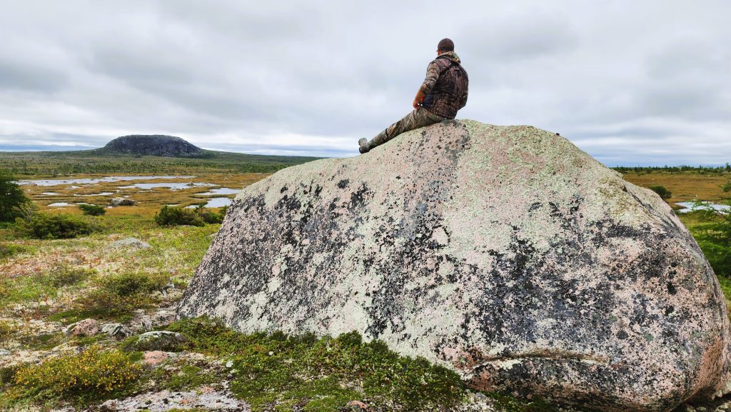 A hunter dressed in camouflage rests on a huge rock while he surveys the landscape for big game, and enjoys the breathtaking beauty of the Newfoundland wilderness