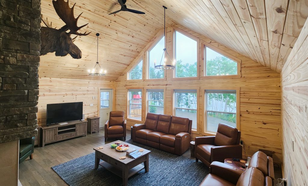Comfortable lodge living room with fireplace, leather furniture, big screen TV and beautiful wilderness views. A large moose head is mounted above the fireplace overlooking the room.