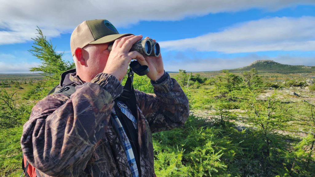 A man holds a set of binoculars up to his eyes as he searches the surrounding Newfoundland wilderness in search of big game.