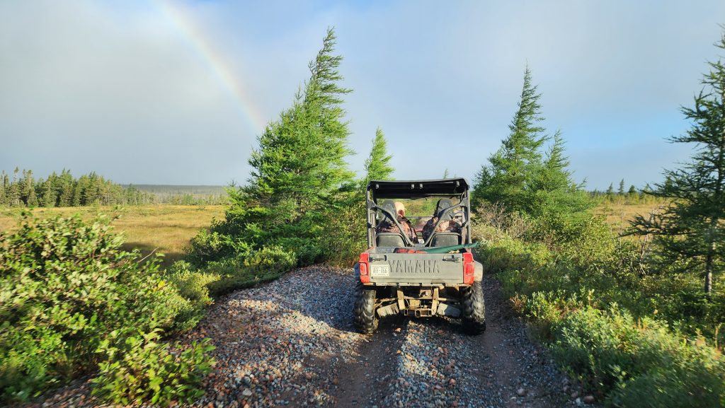 Side-by-side ATV with two hunters crossing rugged Newfoundland terrain with vibrant rainbow in the background