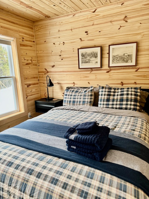 Cozy Newfoundland Adventures hunting lodge room accommodation with queen size bed and scenic window view. The bed is made with a plaid comforter and a stack of folded fresh towels rests on the corner ready for guests to use.