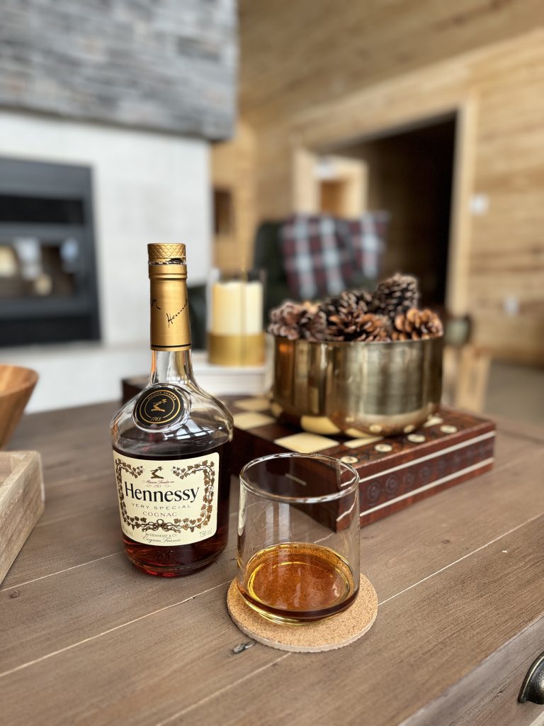 A glass of brandy sits on a coaster beside the bottle of brandy that is being shared by hunters in the luxurious hunting lodge. A bowl of decorative pine cones rests behind it to complete the nature inspired decor of the lodge.